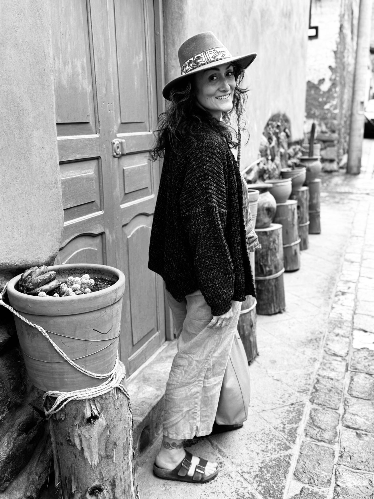 Woman wearing a traditional Andean hat standing in a doorway on a Pisac street, surrounded by adobe walls and plant pots.